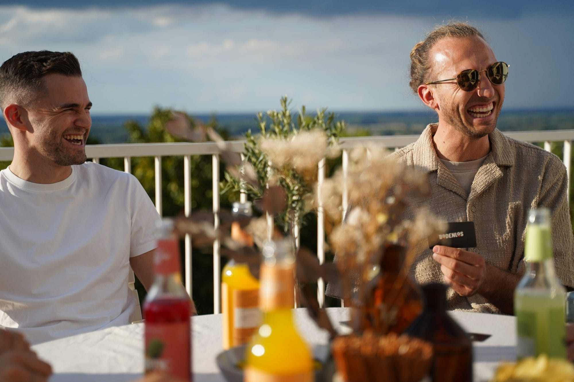 Zwei junge Männer lachen an einem gedeckten Tisch auf einer Terrasse, einer hält eine BODENLOS-Karte.