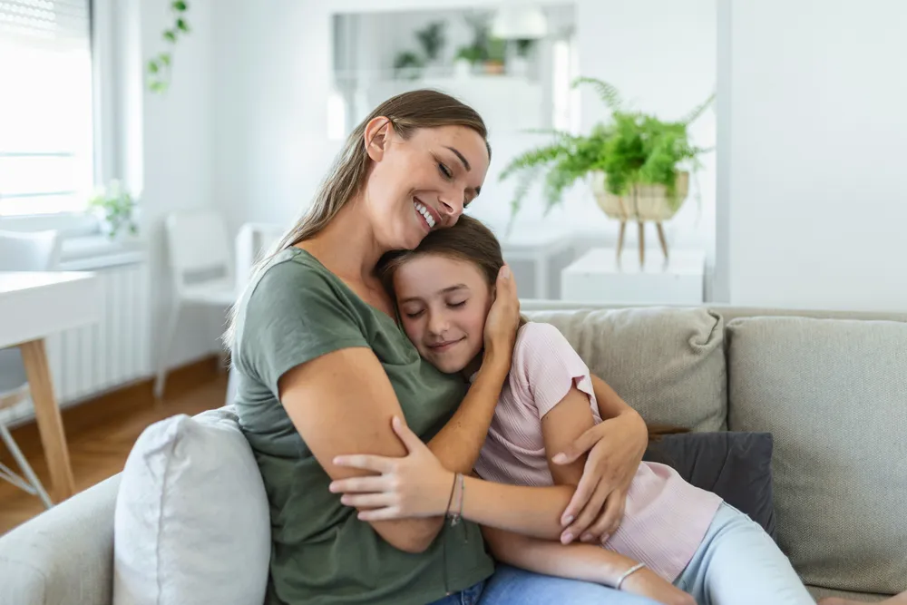 Eine Mutter umarmt ihre Tochter im Teenageralter liebevoll auf dem Sofa.