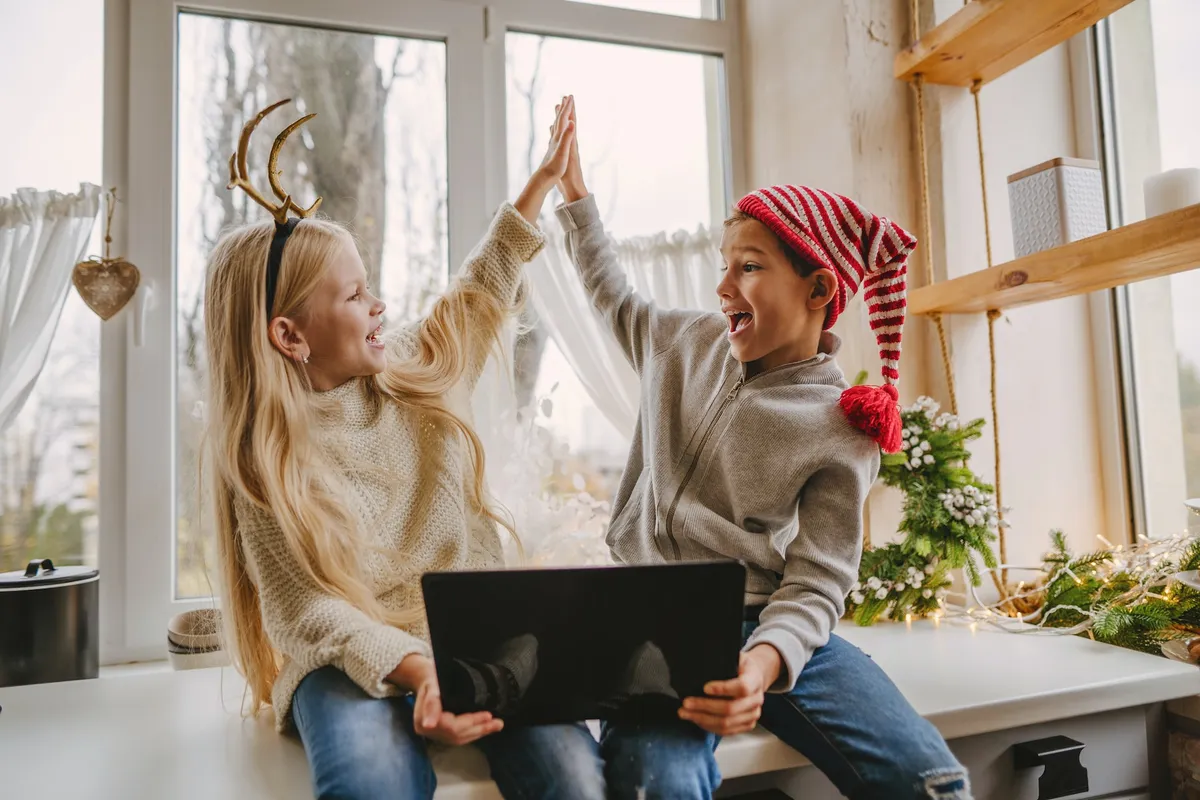 Zwei lachende Kinder mit Rentier-Geweih und Weihnachtsmütze geben sich ein High-Five bei einer Weihnachts-Schnitzeljagd am Laptop.