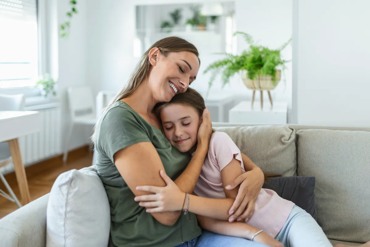 Eine Mutter umarmt ihre Tochter im Teenageralter liebevoll auf dem Sofa.