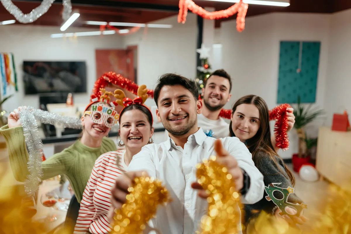Fünf lachende junge Erwachsene mit Lametta, Rentiergeweih und Partybrillen auf einer Weihnachtsfeier