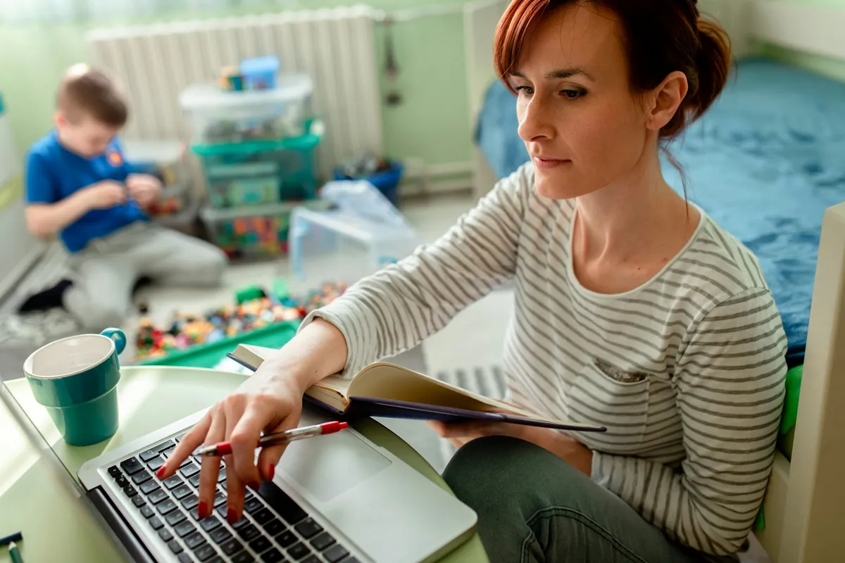 Mutter arbeitet am Laptop und hält ein Notizbuch, während ihr Sohn im Hintergrund auf dem Boden spielt