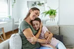Eine Mutter umarmt ihre Tochter im Teenageralter liebevoll auf dem Sofa.