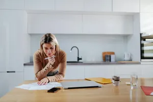 Frau stützt sich in der Küche auf die Arbeitsplatte und geht mit dem Stift in der Hand Rechnungen durch