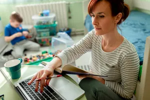 Mutter arbeitet am Laptop und hält ein Notizbuch, während ihr Sohn im Hintergrund auf dem Boden spielt