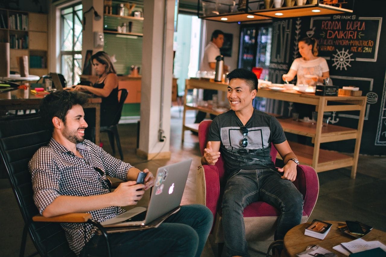 Two young men laughing together in a café, one with a laptop on his lap