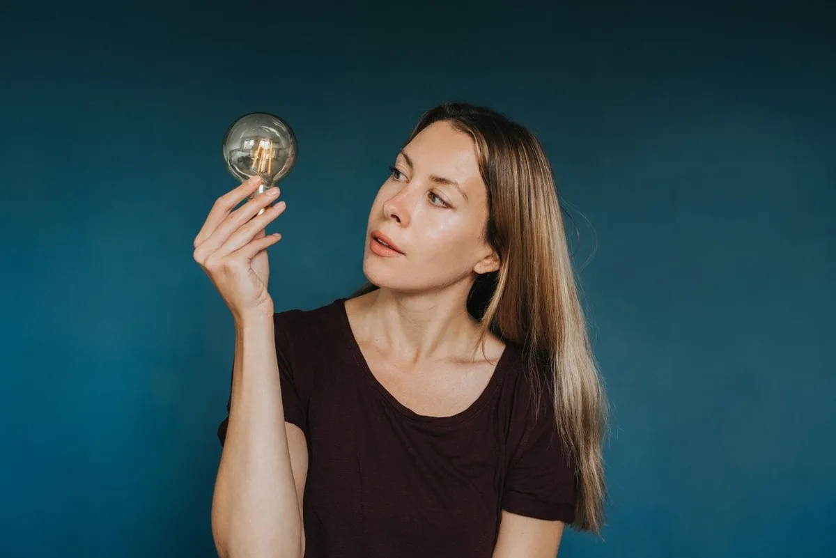 A young woman holds up a light bulb and looks at it, in front of a dark blue wall