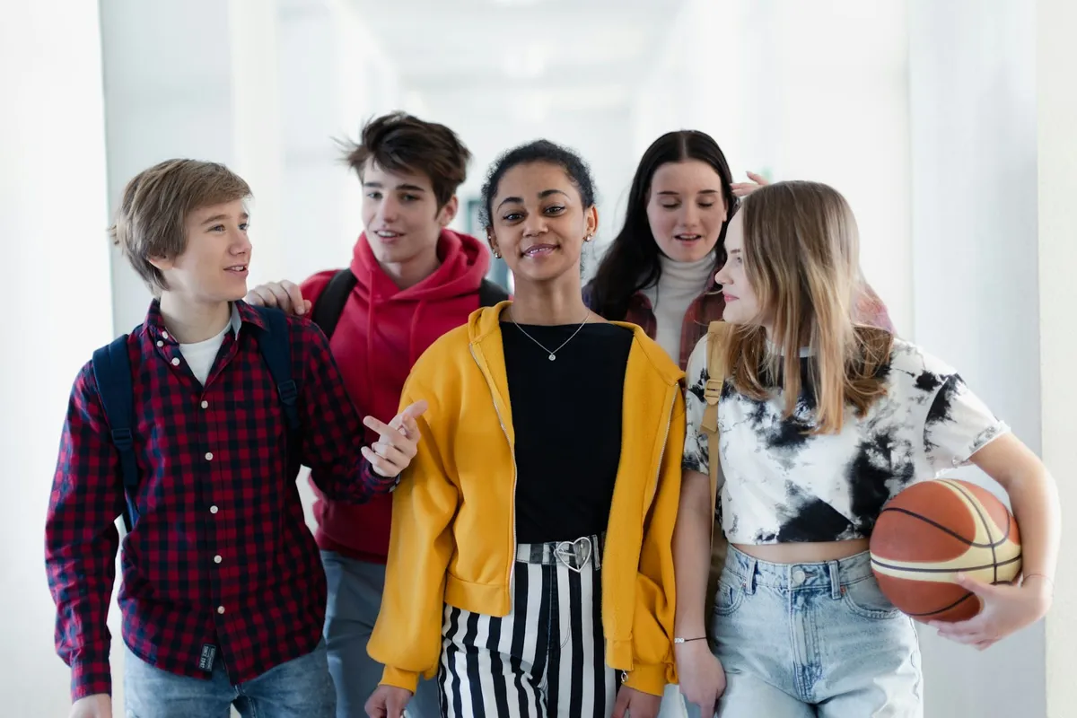 Five teenagers with backpacks walk and talk in a bright school hallway, one of them holding a basketball