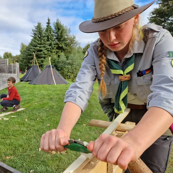 A scout in a hat and green neckerchief smooths a wooden board with a two-handled tool while a boy watches, with black tents in the background