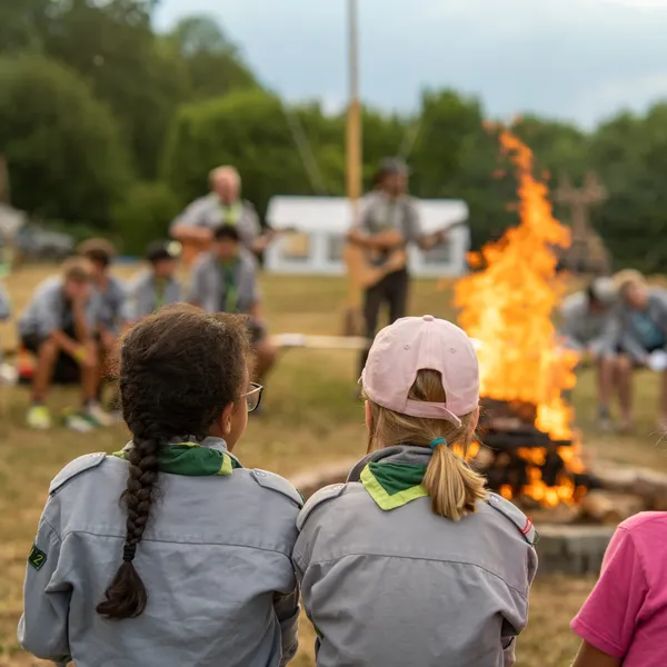 Two girls in gray scout shirts with green neckerchiefs watch a large campfire, while other scouts sit in a circle and two leaders play guitar in the background
