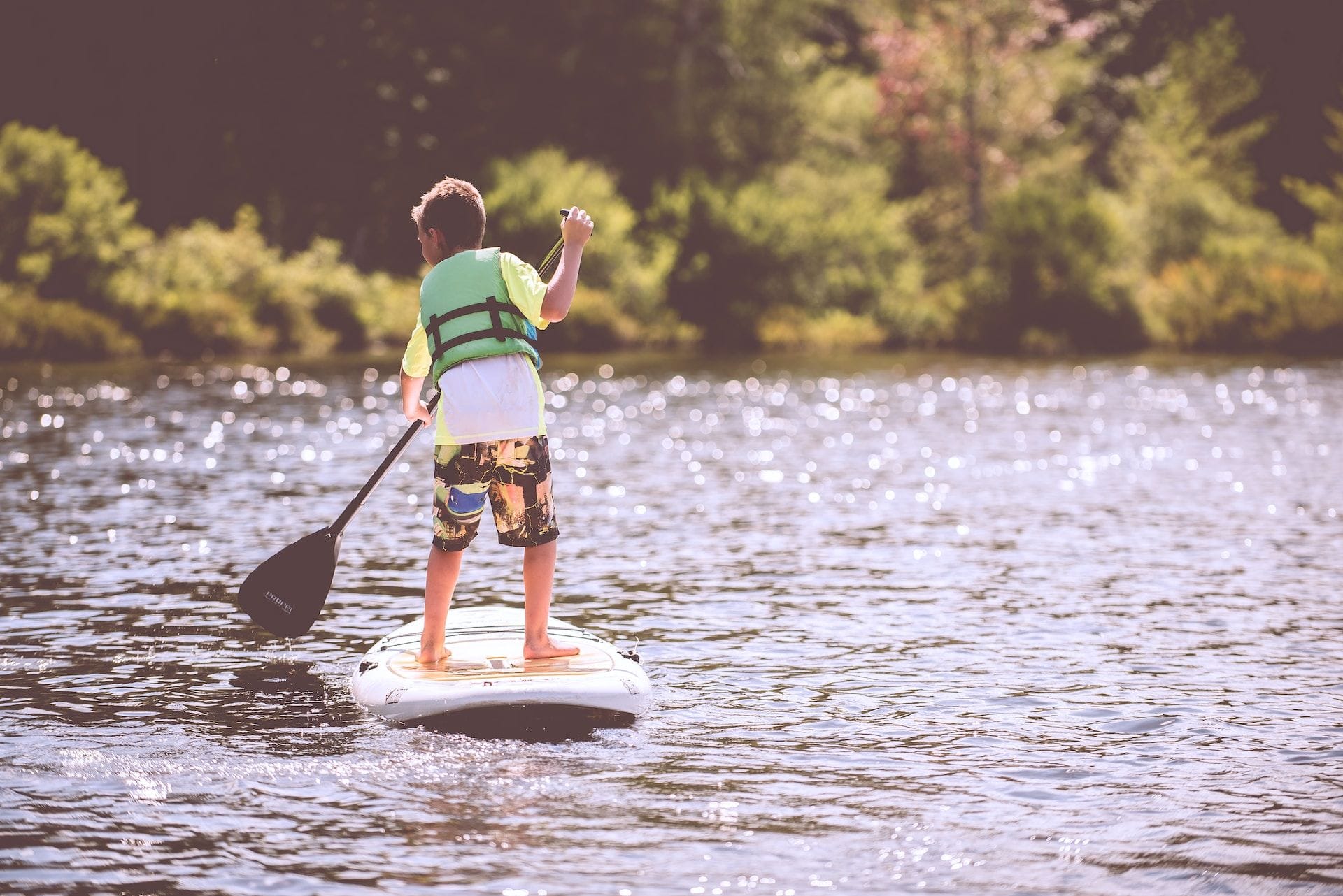 Un niño con chaleco salvavidas rema de pie sobre una tabla en un lago