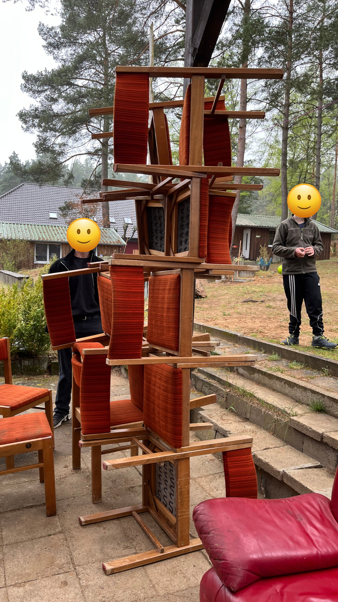 Una torre alta de sillas de madera apiladas al aire libre; al lado, dos jóvenes con la cara tapada por un emoji