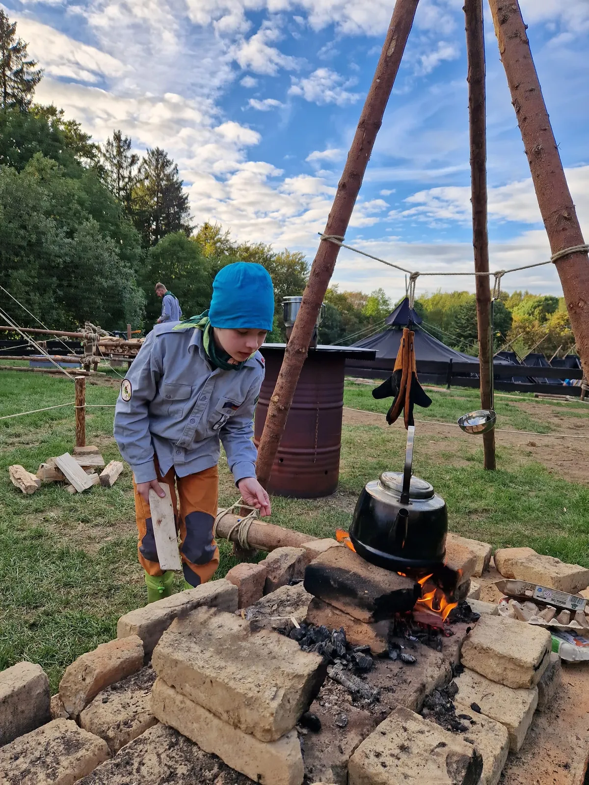 Un scout con gorro azul y pañoleta verde sostiene un trozo de madera junto a un fogón de ladrillos con una tetera negra; al fondo, tiendas negras de scouts