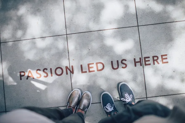 Los pies de dos personas sobre un suelo de baldosas con la frase «passion led us here» pintada