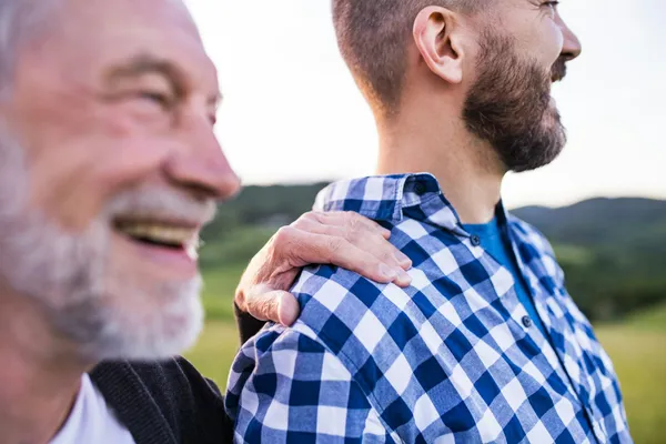 Un hombre mayor sonriente apoya la mano en el hombro de un hombre más joven con barba y camisa de cuadros, al aire libre