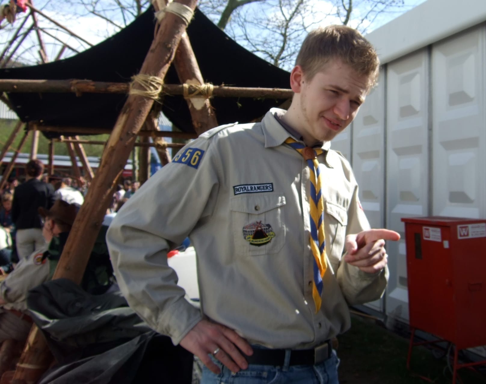 Un jeune homme en chemise scoute beige avec l’insigne des Royal Rangers et un foulard rayé, devant une construction en bois et une tente noire