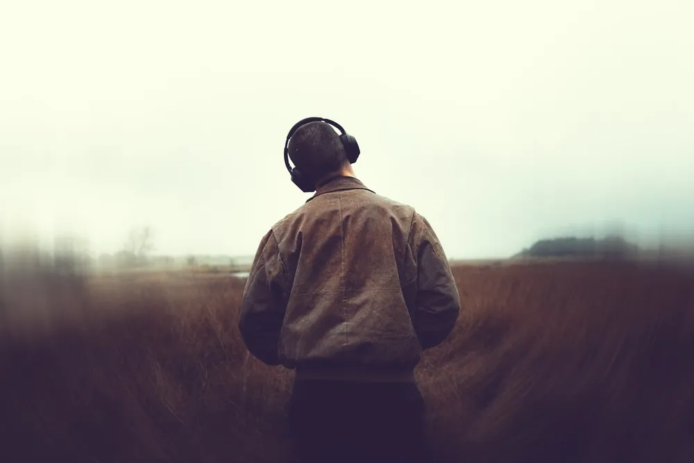 Un homme avec un casque audio, vu de dos, debout dans un champ d’herbes hautes sous un ciel brumeux