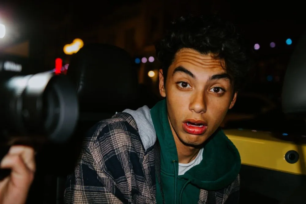 Un jeune homme aux cheveux bouclés, l’air surpris, la nuit, face à un appareil photo