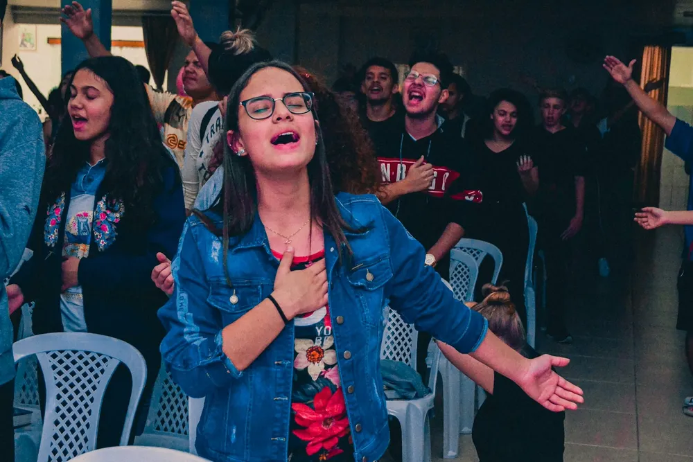 Une jeune fille en veste en jean, lunettes sur le nez, chante les yeux fermés, la main sur le cœur, entourée d’autres jeunes en pleine louange