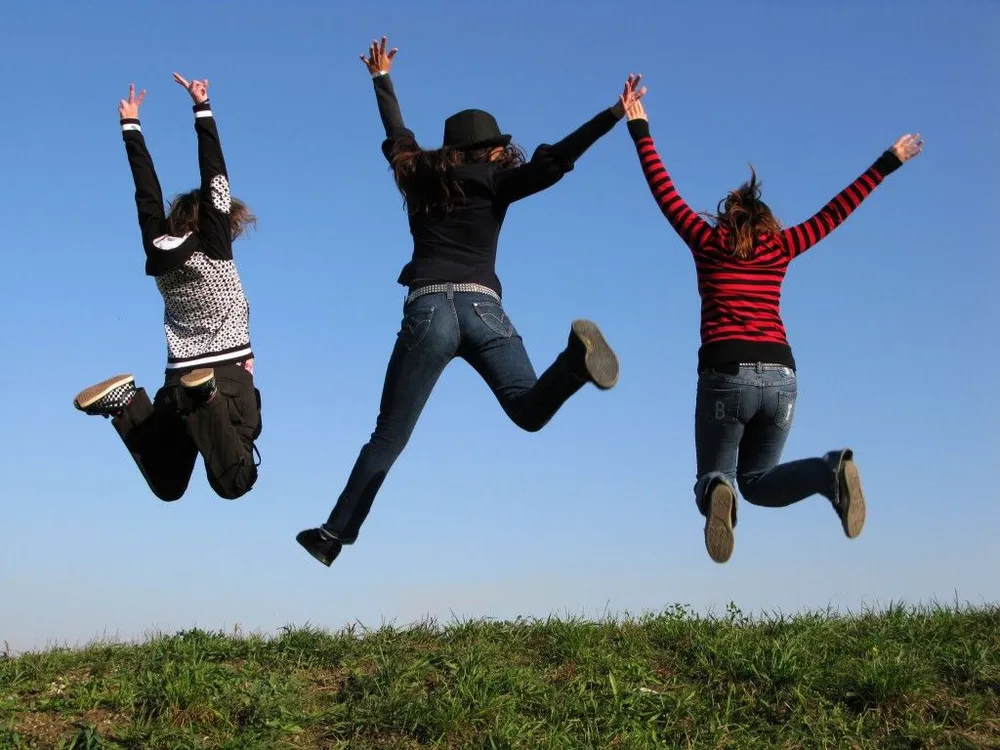 Trois jeunes, vus de dos, sautent bras levés au-dessus d’une prairie, sous un ciel bleu