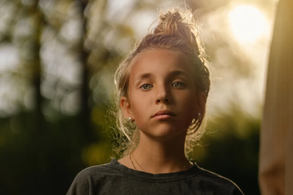 Une jeune fille blonde aux cheveux attachés regarde l’objectif d’un air pensif, en extérieur