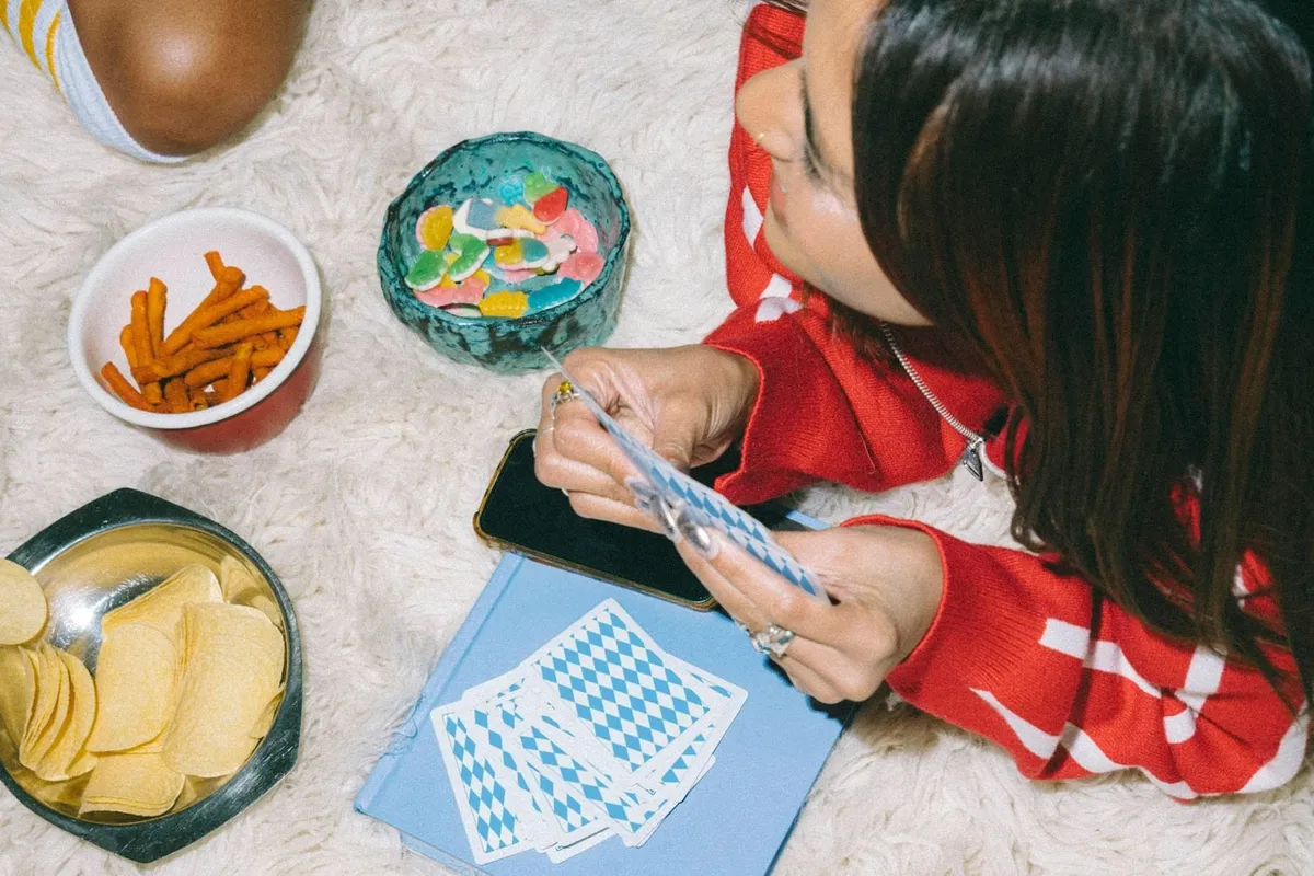 Vue de dessus d’une jeune femme qui joue aux cartes, allongée sur un tapis, entourée de bols de bonbons et de chips