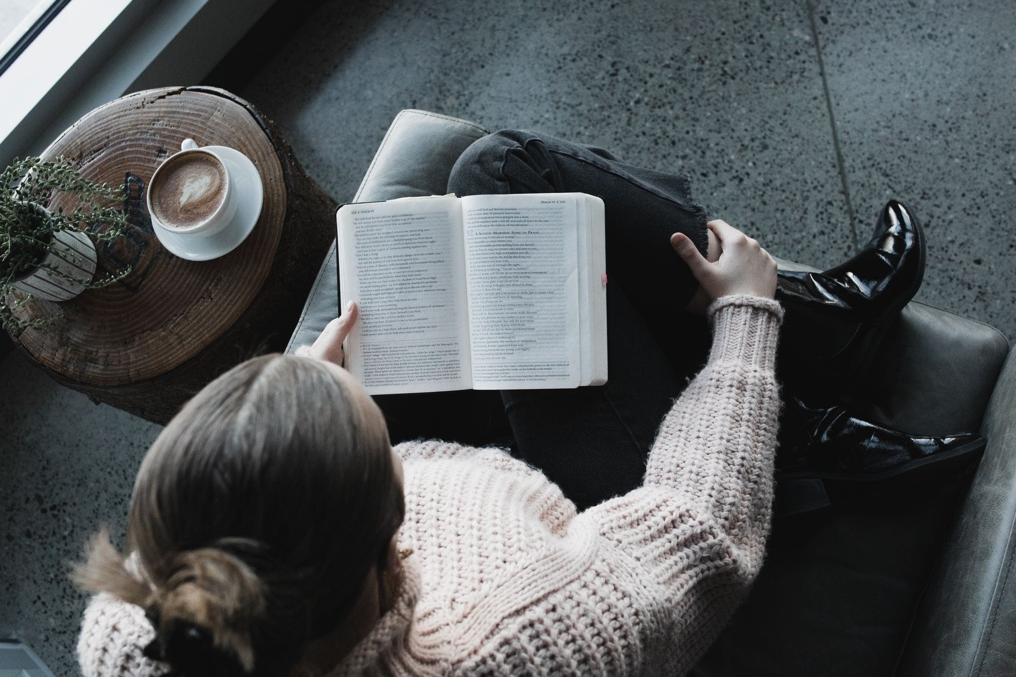 Văzută de sus, o tânără într-un pulover gros citește Biblia într-un fotoliu; lângă ea, pe o măsuță de lemn, o cafea și o plantă