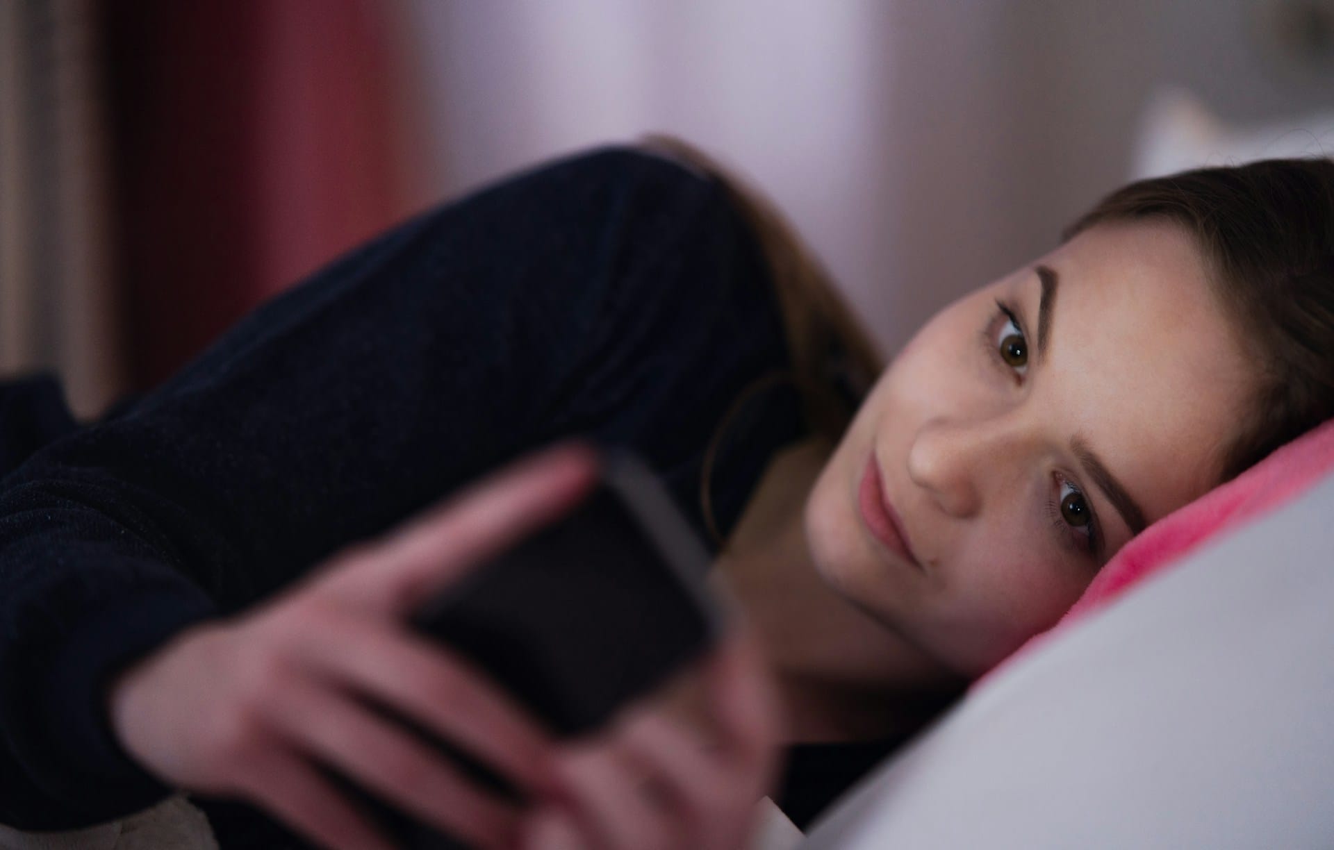 O adolescentă stă întinsă în pat, în semiîntuneric, și se uită la telefonul din mână