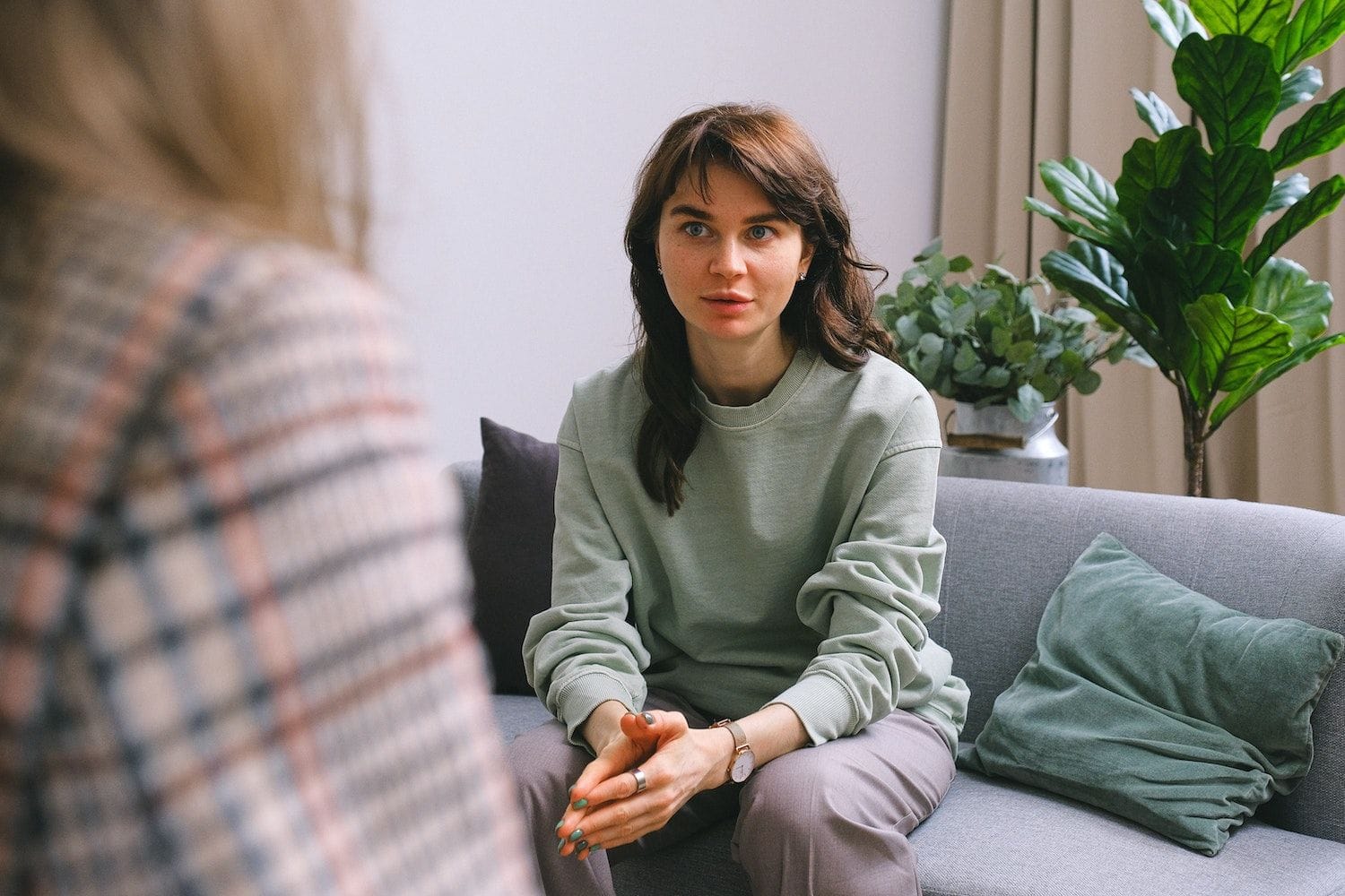 O tânără stă pe o canapea gri și ascultă atentă o femeie care îi vorbește și care se vede din spate
