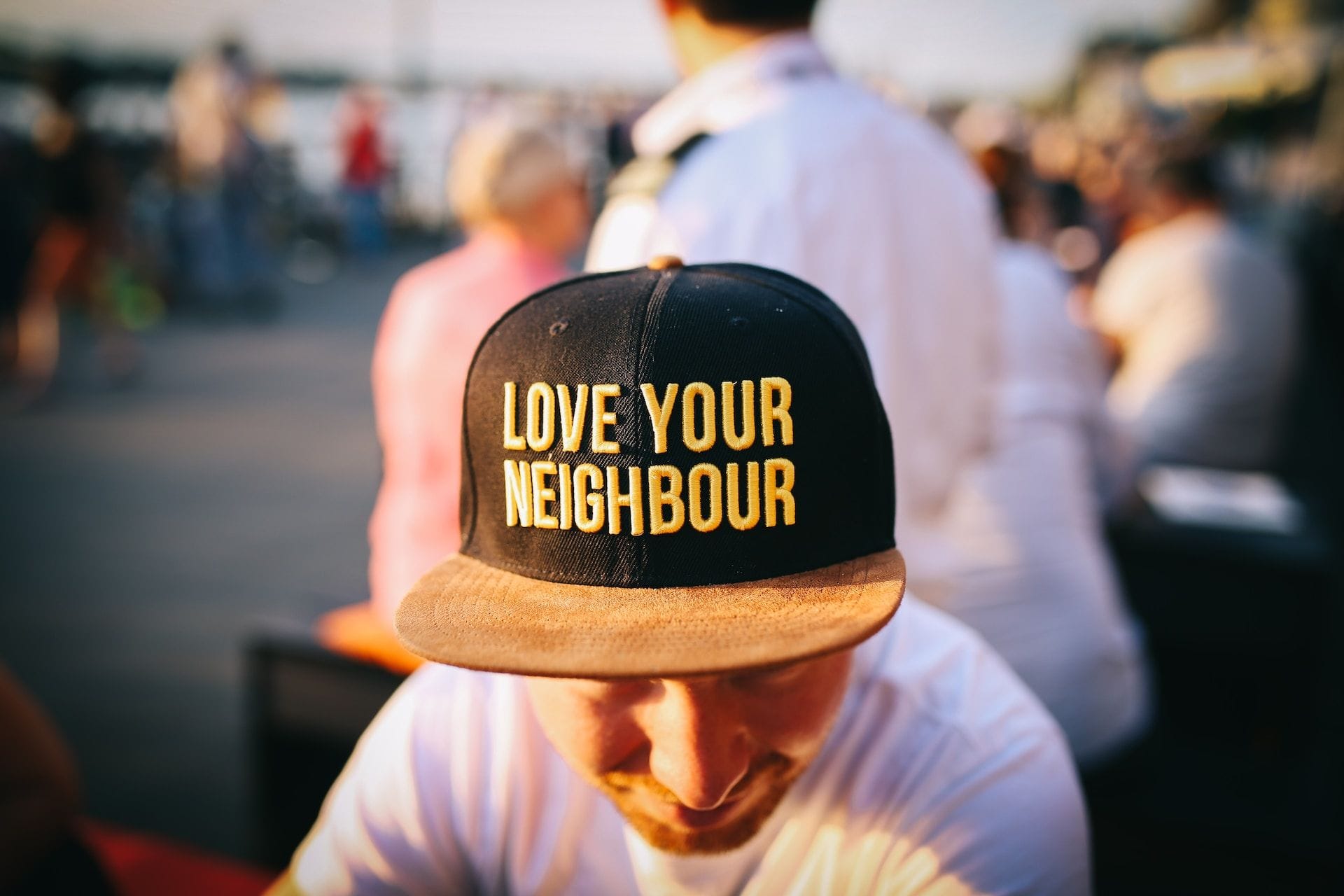 Un tânăr cu privirea în jos poartă o șapcă neagră pe care scrie „Love your neighbour”, în mijlocul unei mulțimi, în aer liber