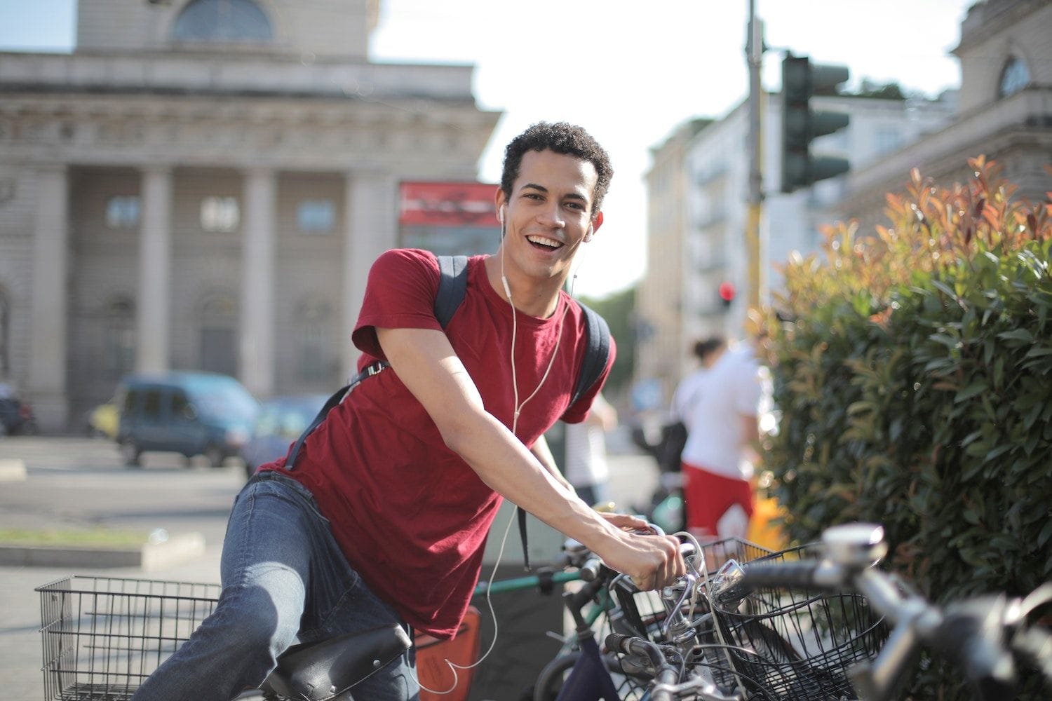 Un tânăr zâmbitor, cu tricou roșu, rucsac și căști, stă pe bicicletă pe o stradă din oraș