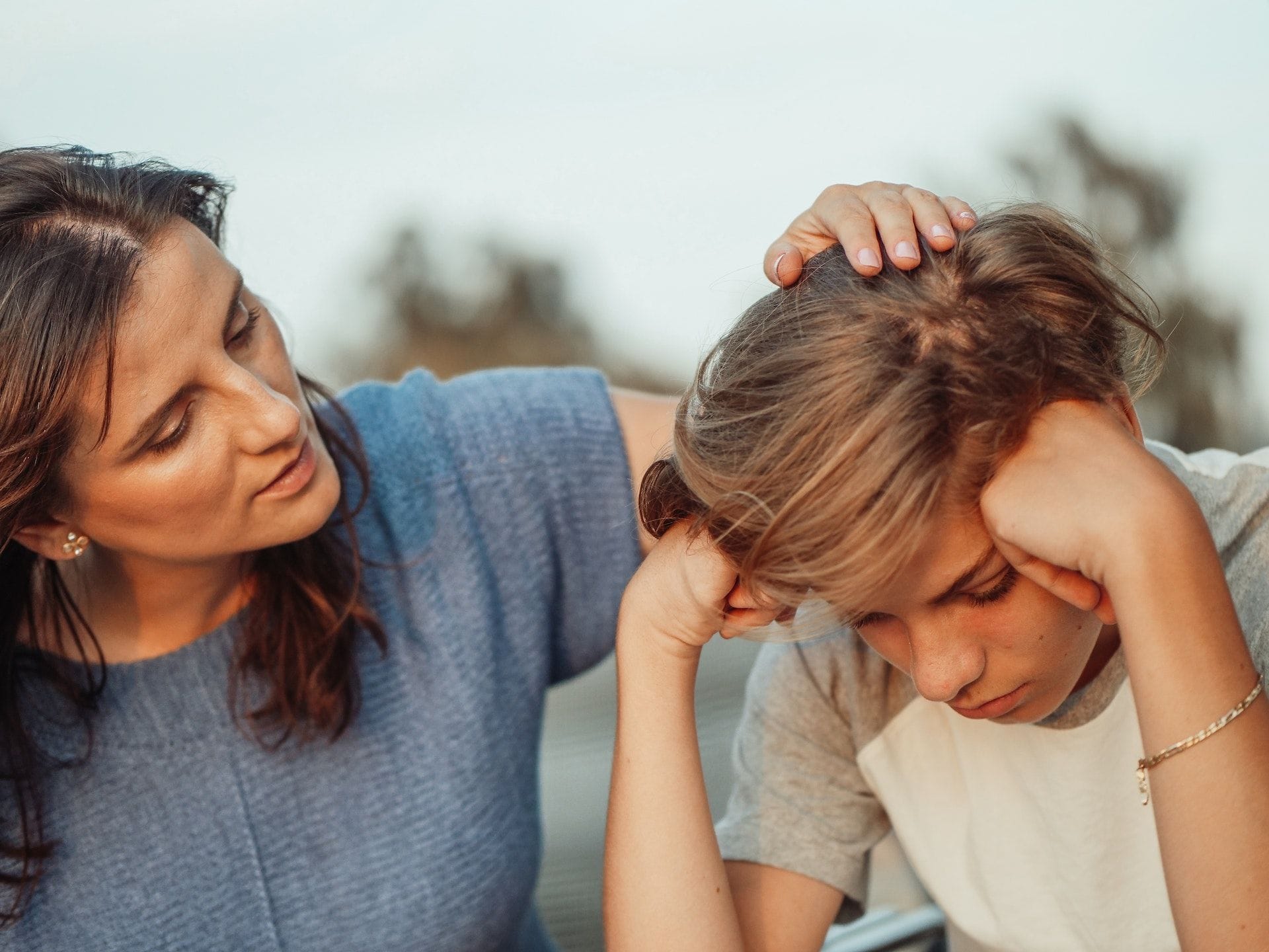 O mamă își pune mâna pe capul fiului ei adolescent, care stă abătut, cu capul sprijinit în mâini