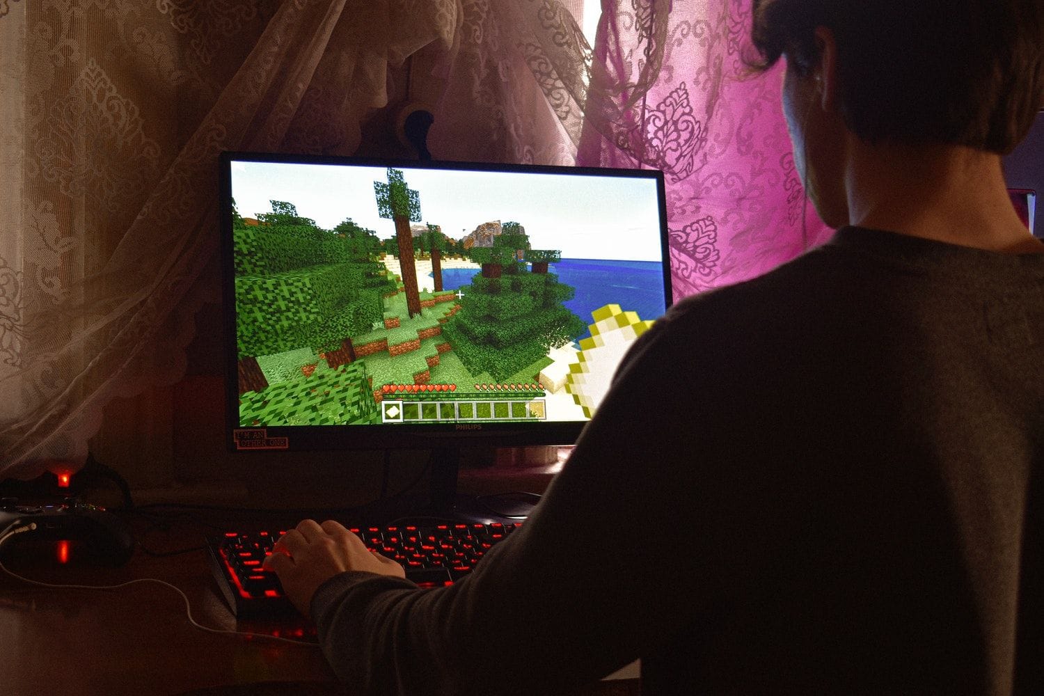 O persoană joacă Minecraft la calculator, cu tastatura iluminată în roșu, într-o cameră cu perdele de dantelă