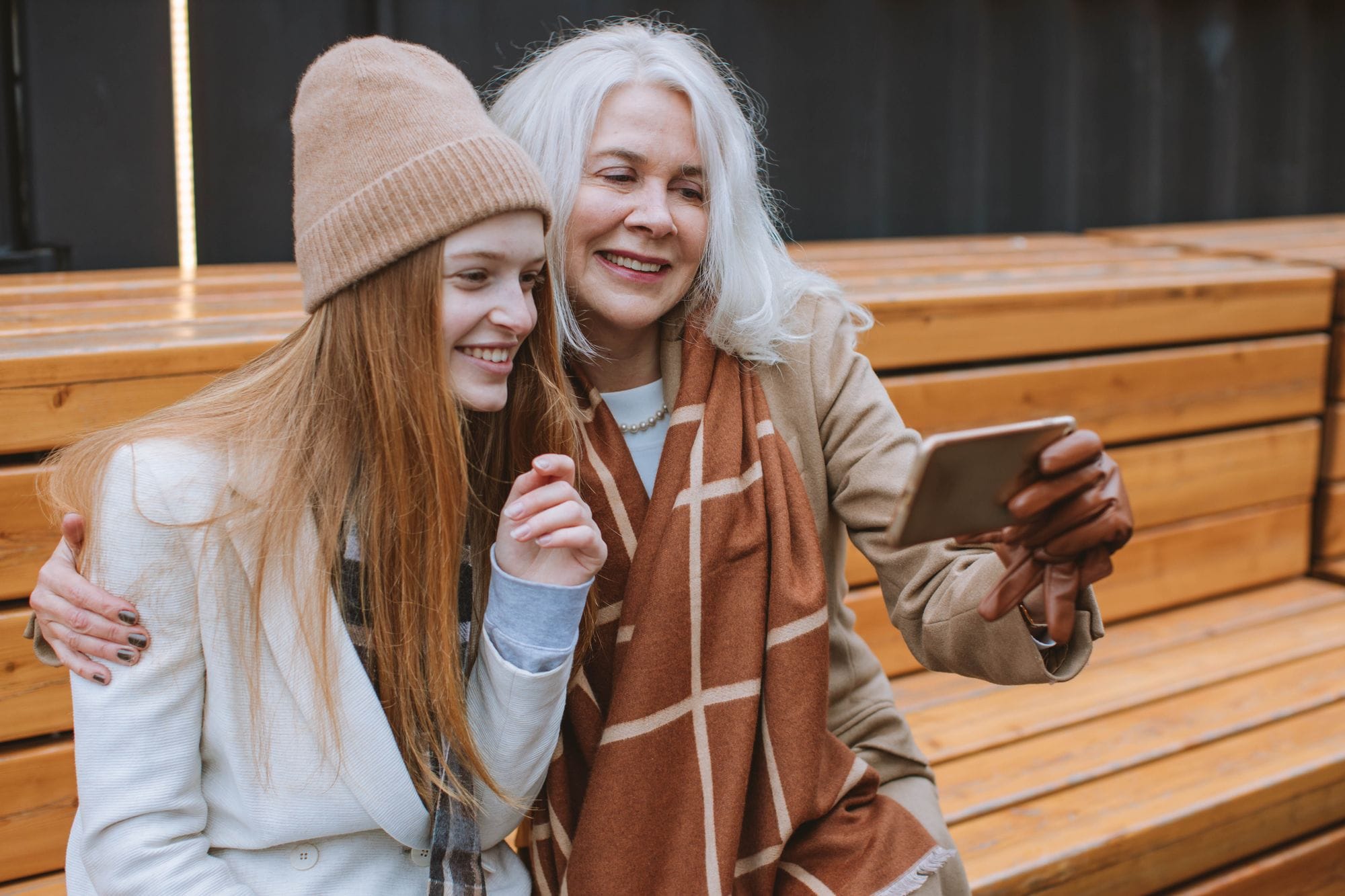 O adolescentă cu căciulă și o femeie cu părul alb își fac un selfie zâmbind, pe o bancă de lemn