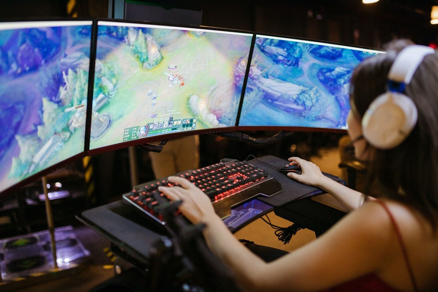 O tânără cu căști joacă League of Legends pe trei monitoare curbate, cu o tastatură iluminată în roșu