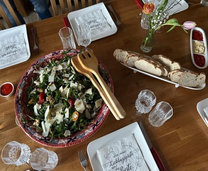 Masă de lemn pregătită, cu un bol mare de salată de rucola, pâine feliată, pahare și flori, la un Running Dinner