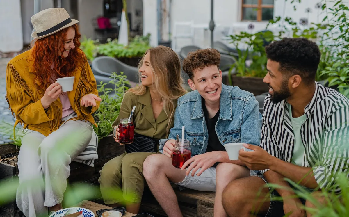 Patru tineri stau pe o bancă în aer liber, între plante, la o cafenea, beau ceai și limonadă și râd împreună