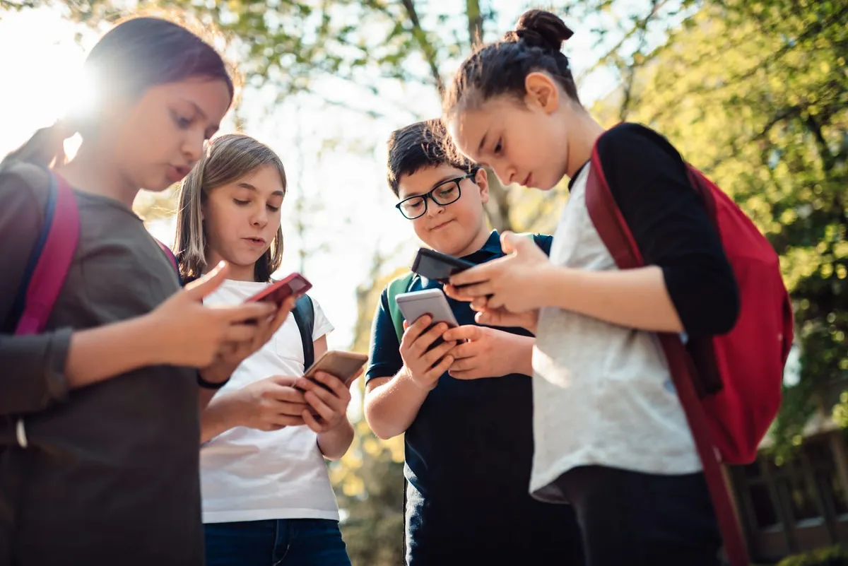 Patru preadolescenți cu ghiozdane, în aer liber, fiecare cu privirea în telefonul lui