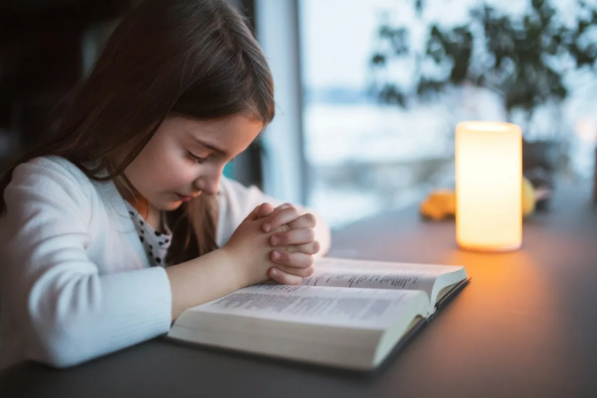 O fetiță se roagă cu mâinile împreunate peste o Biblie deschisă, lângă o lumânare aprinsă