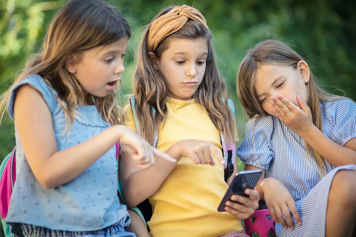 Trei fete se uită uimite la un smartphone; două arată spre ecran, a treia își acoperă gura cu mâna