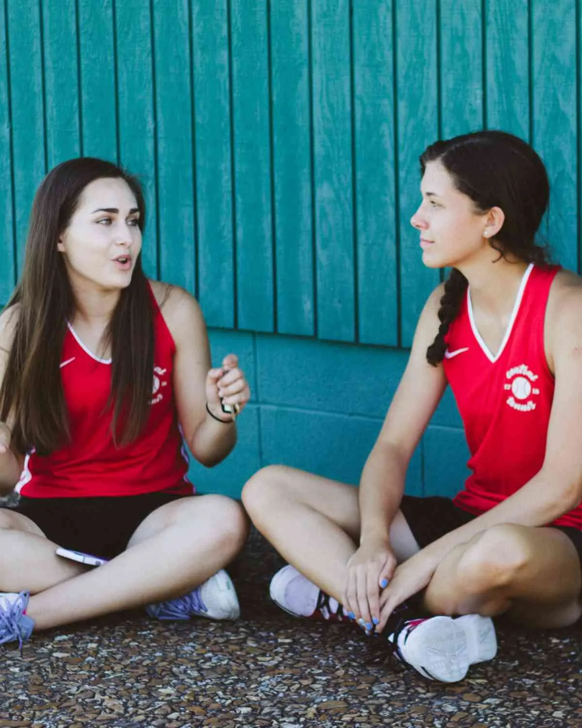 Două adolescente în maiouri sportive roșii stau turcește pe jos, în fața unui perete turcoaz din lemn, și vorbesc