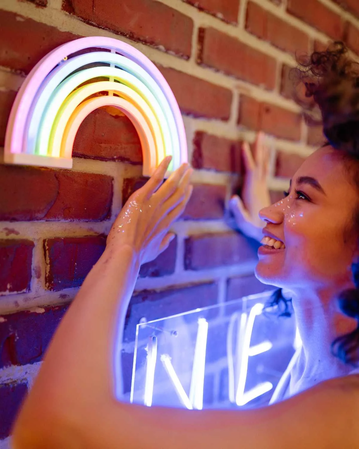 O tânără zâmbește și atinge un curcubeu de neon prins pe un zid de cărămidă