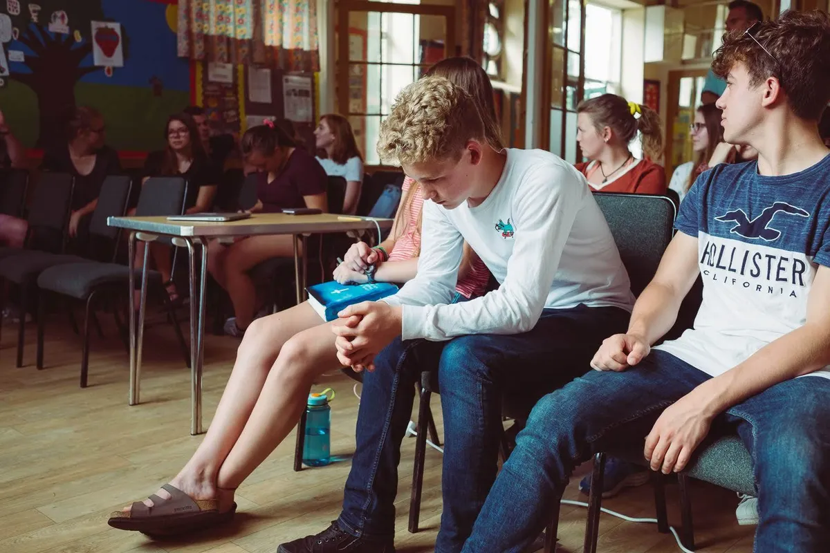 Adolescenți așezați pe scaune într-o sală de întâlnire; un băiat stă cu capul plecat și cu mâinile împreunate, lângă o fată care ține o Biblie albastră