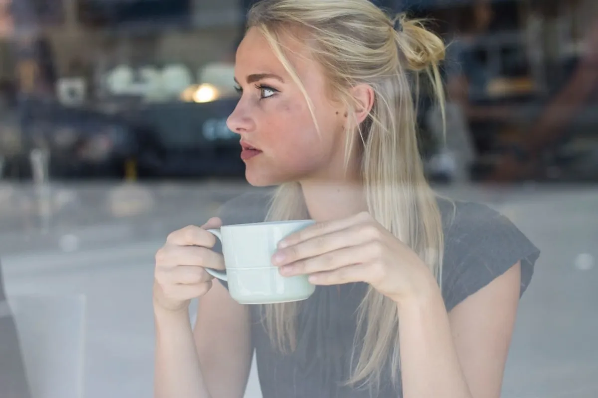 O tânără blondă ține o ceașcă în mâini și privește pe geamul unei cafenele