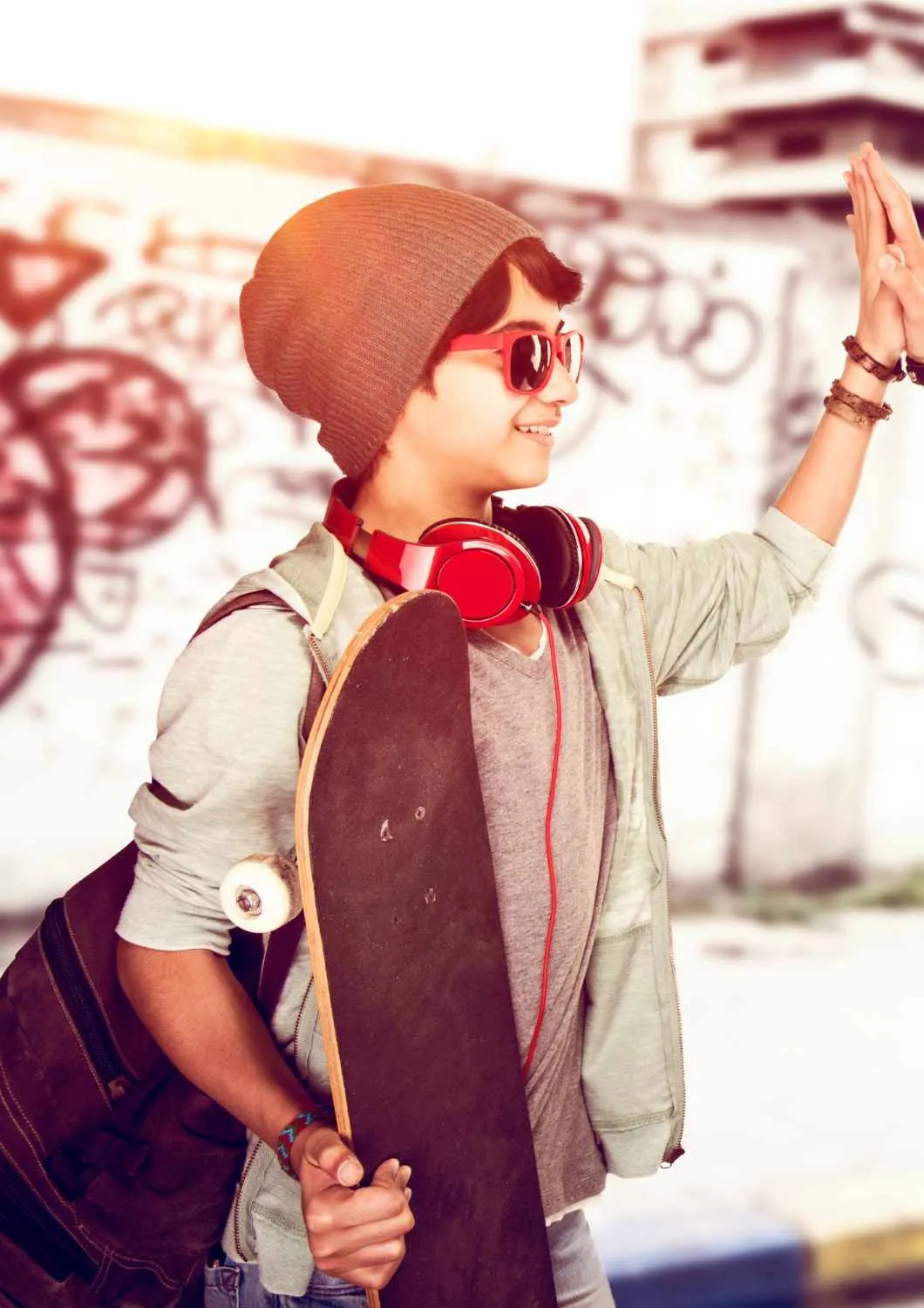 Un adolescent cu căciulă, ochelari de soare roșii și căști roșii la gât ține un skateboard și bate palma cu cineva, în fața unui zid cu graffiti