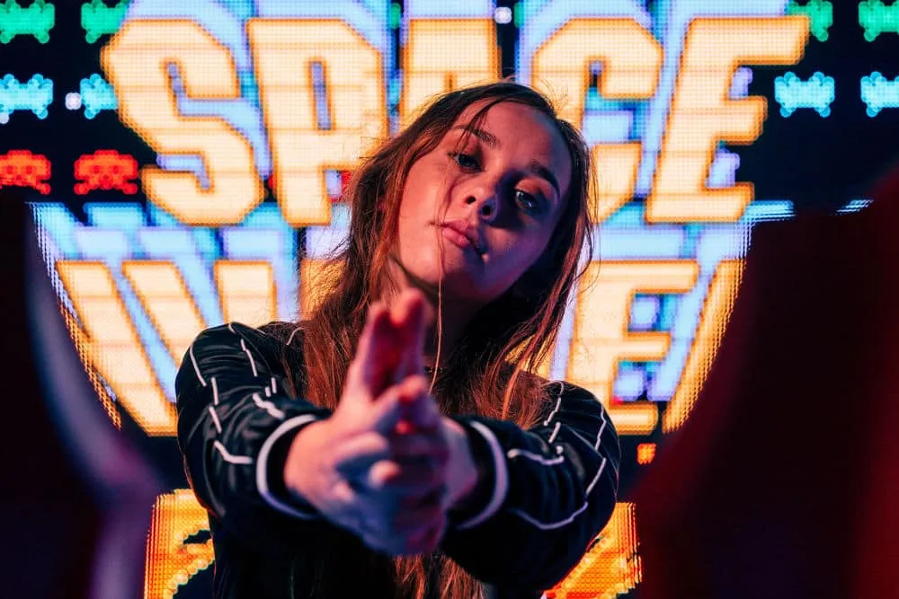 O tânără face cu mâinile semnul unui pistol îndreptat spre cameră, în fața unui ecran luminos cu jocul Space Invaders