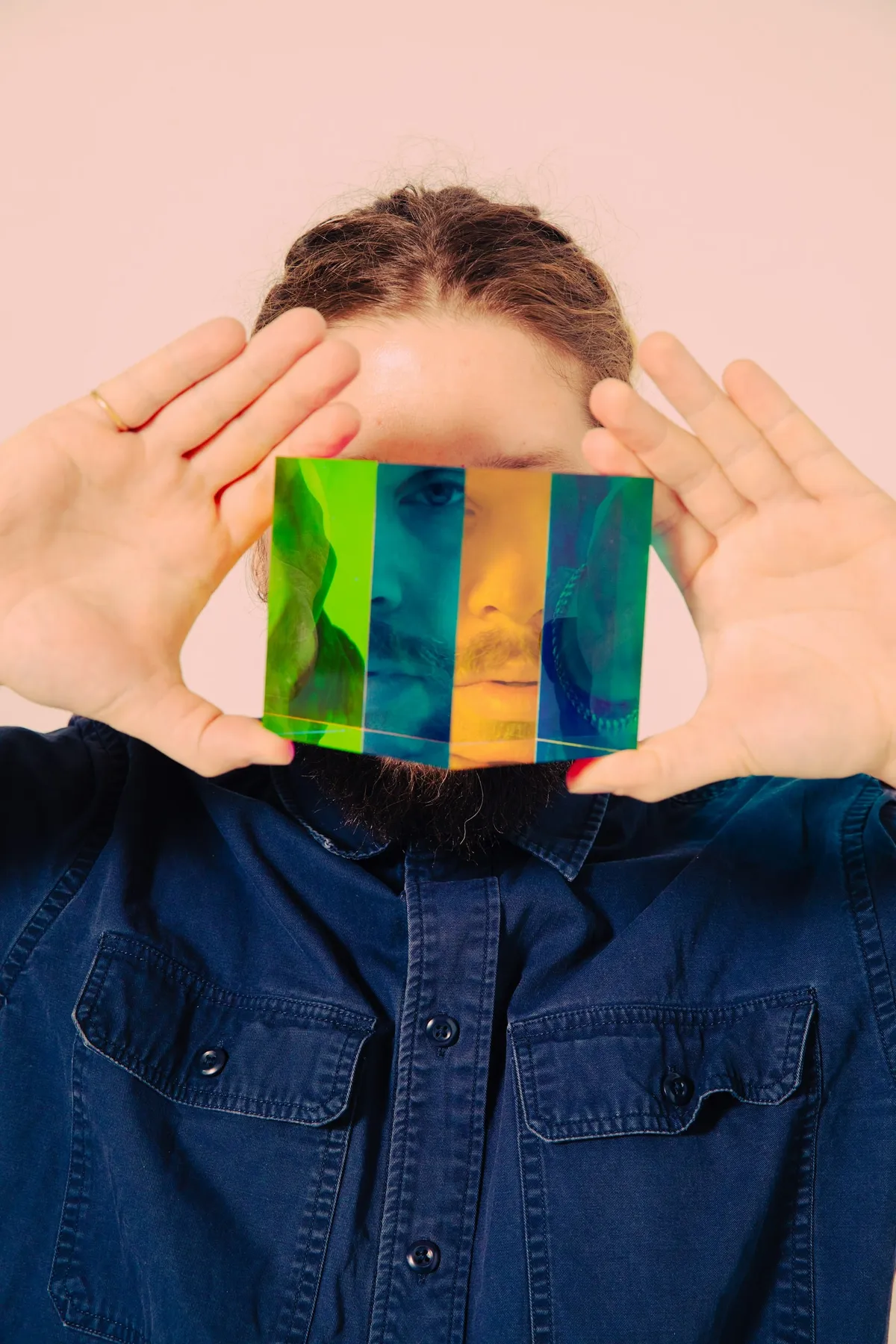 Un bărbat cu barbă și cămașă de blugi ține în fața feței o prismă colorată, prin care chipul lui apare împărțit în fâșii verzi, albastre și portocalii