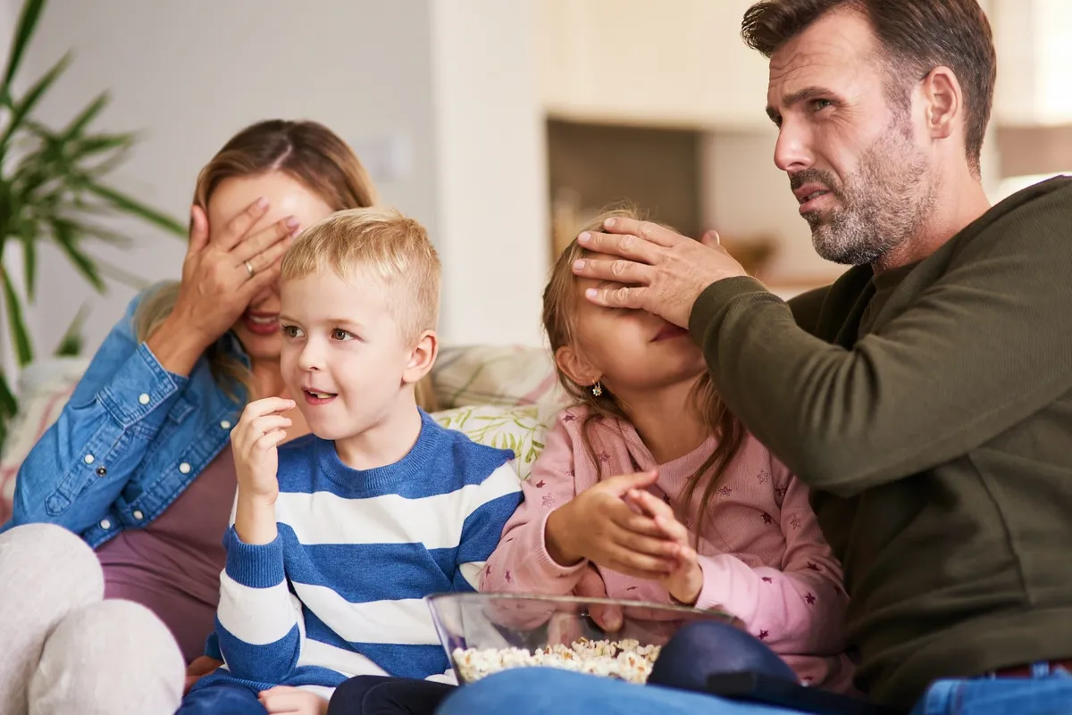 O familie pe canapea se uită la un film: mama își acoperă ochii, tatăl îi acoperă ochii fetiței, iar băiatul mănâncă popcorn și se uită mai departe