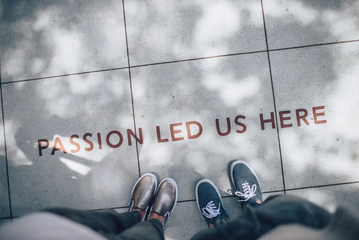 Picioarele a două persoane pe un trotuar, lângă cuvintele în engleză „passion led us here”, pictate pe dale