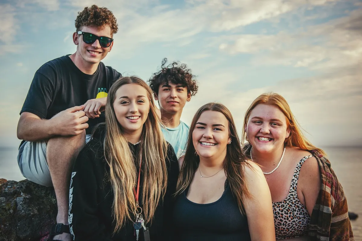 Cinci adolescenți zâmbitori, doi băieți și trei fete, pe o stâncă la malul apei, sub un cer cu nori